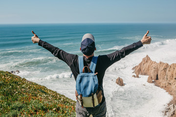 Wall Mural - A tourist with a backpack on a cliff against the background of the Atlantic Ocean raised his hands and shows how he is free and happy.