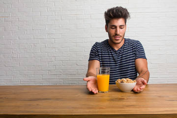 Wall Mural - Young handsome and natural man sitting on a table holding something with hands, showing a product, smiling and cheerful, offering an imaginary object. Having a breakfast