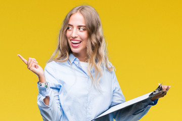 Sticker - Beautiful young blonde business woman holding clipboard over isolated background very happy pointing with hand and finger to the side