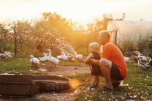 Grandfather Playing With His Grandson While Crouching And Spraying With Hose. In Background Ducks And Hens. Countryside Exterior.