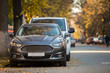Front view of gray expensive luxurious car parked in quiet alley on sunny autumn day on blurred vehicles, walking people and golden foliage bokeh background. Transportation, parking problems concept.
