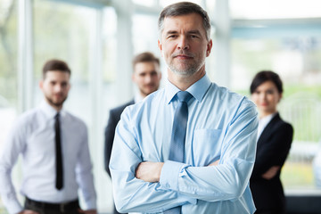 Wall Mural - Modern mature businessman smiling and looking at camera with his colleagues in the background at office.