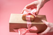© Artem - Hands holding gift box Packed in Kraft paper on pink background. Holiday card for Valentine's day and women's day
