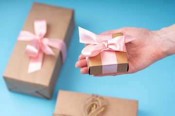 Wall Mural - Women's hands holding gift box Packed in Kraft paper with pink ribbon on blue background. Card for Valentine's day