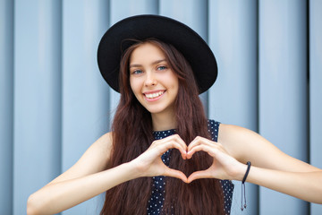 Happy brunette girl in hat making heart sign with her hands at the shutters background