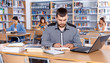 © JackF - Focused guy preparing for exam in college library, reading book and using laptop for searching information
