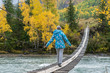 © Евгений Медведев - The girl in a warm jacket goes on a wooden suspension bridge on the other side of the turquoise river. Autumn mountain landscape with yellow fallen leaves