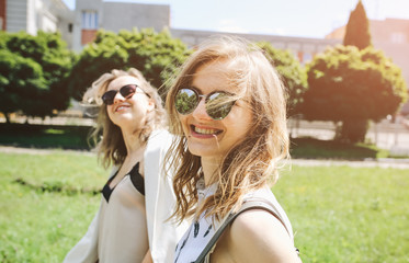 Two stylish hipster girls in glasses are walking in the street. Bright summer photoshoot for women. Beautiful sisters enjoying sun outdoors.