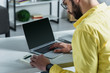 © LIGHTFIELD STUDIOS - bearded man looking at calculator near laptop with blank screen in modern office
