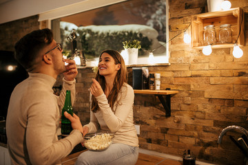 Two young people in bright casual clothing eating popcorn on winter night at home.