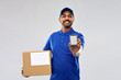 mail service, technology and shipment concept - happy indian delivery man with smartphone and parcel box in blue uniform over grey background