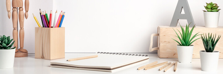 Creative hipster desk by the white wall with wooden accessories. Natural drawing space. Succulents, colored pencils, sketchbook and wooden figure of a man. Panoramic copy space for text