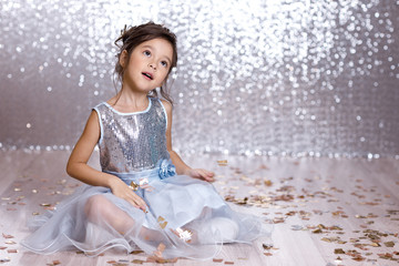 little child girl in blue dress sitting on the floor with confetti on background with silver bokeh. birtday party