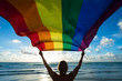 © lazyllama - Scenic silhouette of a man with blond hair holding a gay pride rainbow flag blowing in the wind on a tropical beach with golden sun