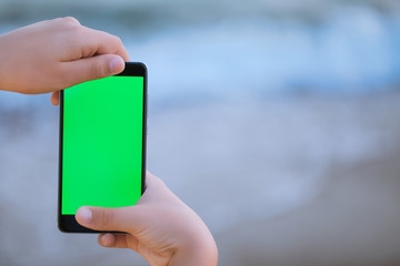 Closeup view of two small hands of young kid holding modern black smartphone with empty blank green screen monitor isolated on blurry blue sea water background. Horizontal photography with copyspace.