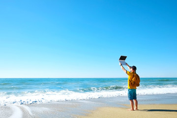 Fit travel blogger holding laptop, writing article on white laptop, standing on empty beach. Freelance remote work concept. Self employed man in yellow t-shirt coding. Copy space, sea view background.