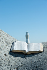 Wall Mural - Open Bible in Psalm 119 on a large stone. Background with blurred white lighthouse in the center of the Bible and a beautiful blue sky. Copy space. Vertical image.