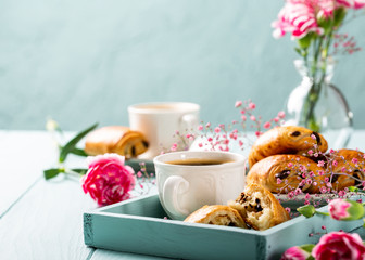 Holiday break with cup of coffee, mini fresh croissants chocolate bun and carnation flowers on turquoise background.