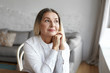 © shurkin_son - Indoor portrait of beautiful 50 year old Caucasian woman with thick loose hair sitting by the window in living room having dreamy pensive look, daydreaming or thinking about something pleasant