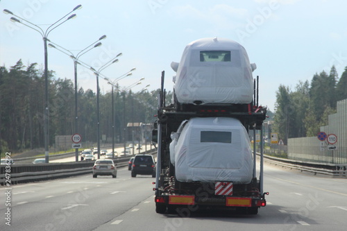 Heavy Car Carrier Truck Transports New Cars In Protective Covers On Heavy Car Carrier Truck Transports New Cars In Protective Covers On Semi Trailer Suburban Asphalt Highway Road Summer Day Against Forest And Blue Sky
