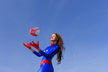 A young woman dressed in shiny blue throws a decorative red heart  shaped gift in the air. She celebrates Valentine's Day and her love to her partner.