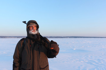 Man with a frozen beard and a tripod on his shoulder on a snowy background in the winter at sunset