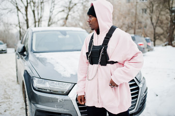 Wall Mural - Stylish urban style african american man in pink hoodie posed against suv car at winter. Afro rapper guy.