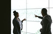 © JonoErasmus - Silhouette of a black african business people giving a presentation conference. Pointing at a large video screen with charts and graphs next to her in a dark room
