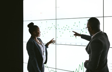 Silhouette of a black african business people giving a presentation conference. Pointing at a large video screen with charts and graphs next to her in a dark room