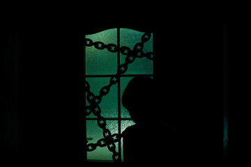 Dark silhouette of kid behind glass door with chain in supernatural green light. Locked alone in room behind door on chain on Halloween. Night kidnapping. Evil in home. Inside haunted house.