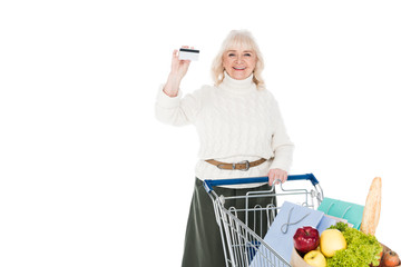 smiling senior woman holding credit card near shopping trolley isolated on white