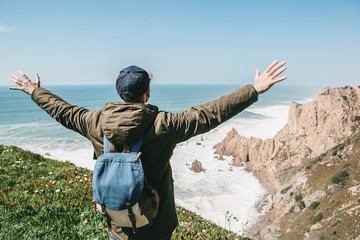 Wall Mural - A tourist with a backpack on a cliff against the background of the Atlantic Ocean raised his hands and shows how he is free and happy.