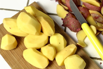 A peeled raw potato, a vegetarian ingredient for cooking meals, on a wooden board next to a potato peel and a knife for peeling potatoes in the kitchen. Dinner preparation process