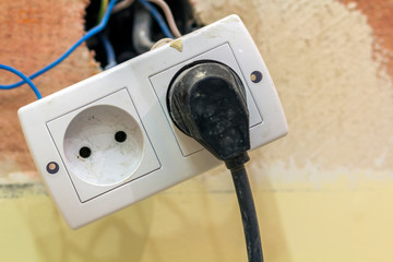 Power outlet in double socket with connecting electric isolated colorful wires, plastic plug installation on light yellow plastered wall. Home interior, electrician job concept.