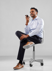 Sticker - business, communication and people concept - smiling indian businessman sitting on office chair using voice command recorder on smartphone over grey background