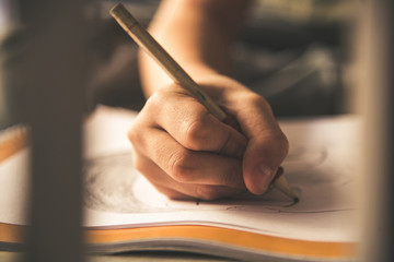 Close up view of a young hand drawing on a white sheet. Kid hold a black wooden pencil and draw something on a warm orange light at home. Children writing on a paper. Teen drawing freehand a manga.
