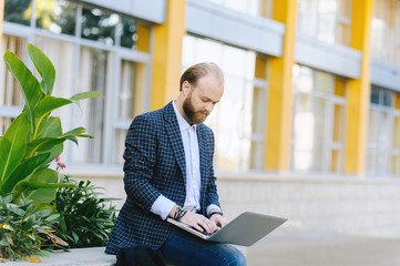 Young bearded businessman sits outdoor on parapet, using laptop and looks on his screen. Social network, checking email.