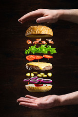 Wall Mural - Floating burger isolated on black wooden background in men hands. Ingredients of a delicious burger with ground beef patty, lettuce, bacon, onions, tomatoes and cucumbers