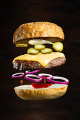 Wall Mural - Floating burger isolated on black wooden background. Ingredients of a delicious cheeseburger with ground beef patty, cheese, onions and cucumbers