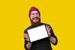 © Vulp - Cheerful bearded man in red hat on yellow background demonstrating white screen of his tablet and smiling at camera
