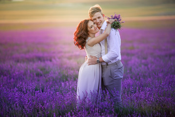 Wall Mural - Couple is hugging and standing on lilac lavender flowers field at blurred background of mountain, nature after wedding ceremony. Bride is dressed in white wedding dress. Bride and groom are married.