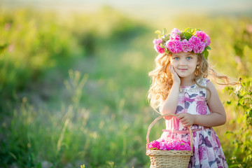 A little girl with beautiful long blond hair, dressed in a light dress and a wreath of real flowers on her head, in the garden of a tea rose