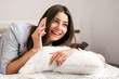 © zadorozhna - Young smiling woman lying in white bed and using a phone in her bedroom. Happy morning.