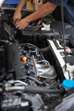 Fototapeta  - Car service by an auto mechanic in garage, fixing a car battery with wrench, also checking some parts of the engine showing by selective focus.