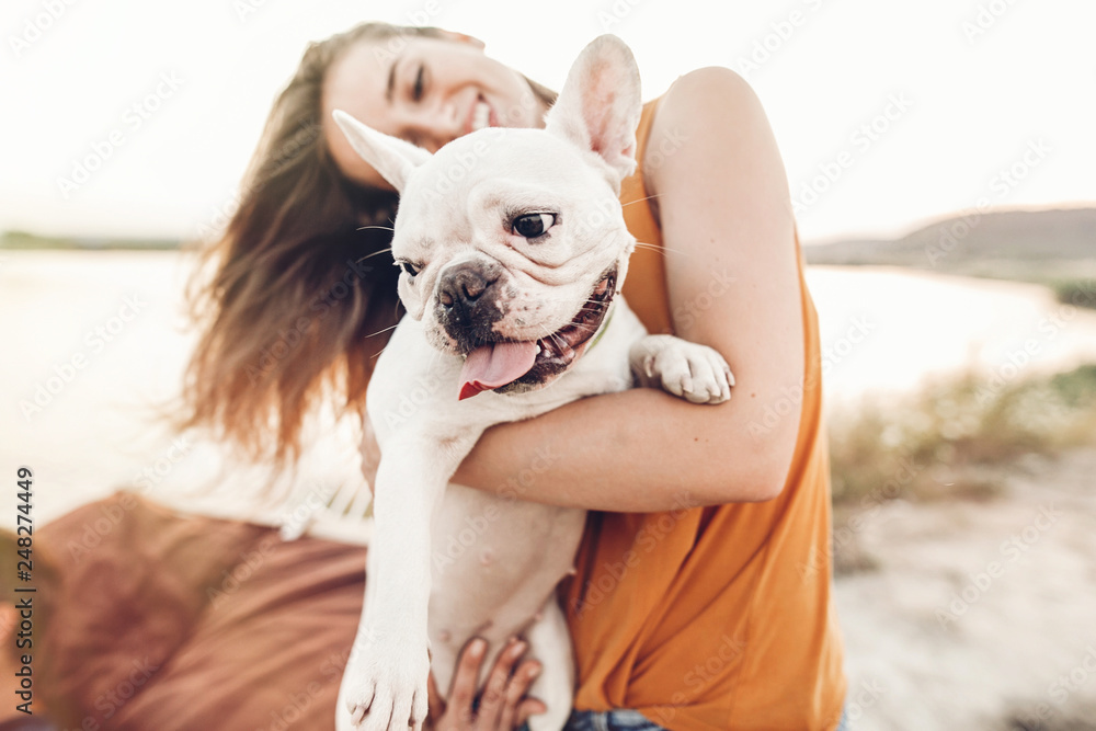 happy hipster woman playing with bulldog on the beach in sunset light, summer vacation. stylish girl with funny dog resting, hugging and having fun in sun, cute moments. space for text - obrazy, fototapety, plakaty 