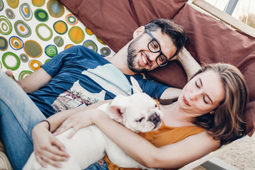 happy hipster couple with bulldog relaxing in hammock on the beach in sunset light, summer vacation. stylish family with dog cuddling and having fun, cute moments in summer evening