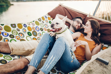 Wall Mural - Cute dog smiling while on a trip with his owners, joyful young family, man and woman lying in comfortable hammock on a beach