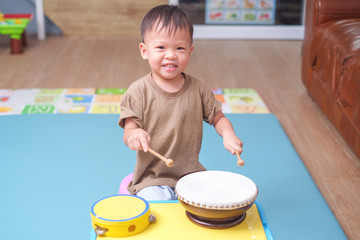 Cute happy smiling little Asian 2 - 3 years old toddler baby boy child hold sticks & plays a musical instrument drum in play room at home, Educational toy for kids and toddlers concept