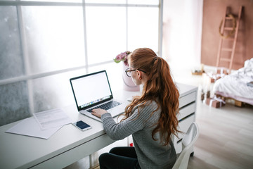 Wall Mural - Young beautiful woman with red hair, wearing glasses, working in the office, uses a laptop and mobile phone