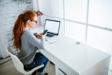 Wall Mural - Young beautiful woman with red hair, wearing glasses, working in the office, uses a laptop and mobile phone